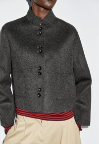 Osoba w ciemnoszarej zapinanej kurtce, pod którą ma na sobie czerwoną koszulkę w paski i beżowe spodnie, z rękami w kieszeniach, na jednolitym białym tle.