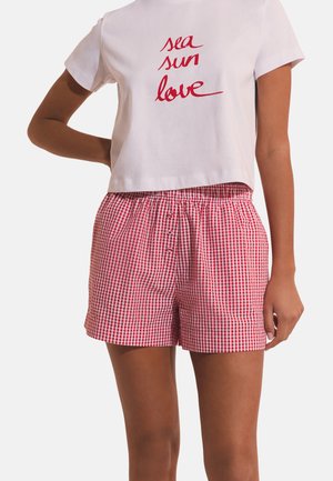 Biała krótka koszulka z czerwonym napisem „sea sun love” w połączeniu z czerwono-białymi, kratkowymi szortami na guziki z przodu na osobie.