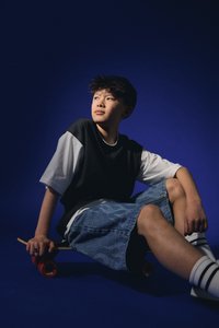 Teenager, oblečený v černobílém oversized tričku a džínových šortkách, sedící na skateboardu s červenými koly, na tmavě modrém pozadí.