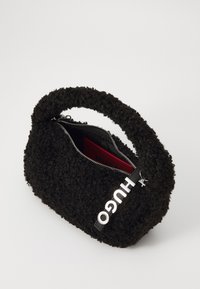 Svart, fluffy handbag med rund form, glidelås og en fremtredende "HUGO"-logo i hvitt. Innvendig har den rødt fôr.