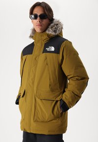 Olivengrøn isoleret jakke med sorte accenter, pelsforet hætte, flere frontlommer og The North Face-logo på brystet.