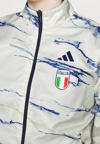 Vit jacka med blå marmormönster, hel dragkedja fram, hög krage och marinblå detaljer. Har Adidas-logotyp och Italiens nationalsymbol.