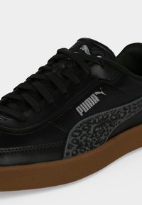 Μαύρο sneaker Puma με μαύρα κορδόνια, λωρίδα με σχέδιο λεοπάρ στα πλευρά, λευκό λογότυπο Puma και καφέ λαστιχένια σόλα σε λευκό φόντο.