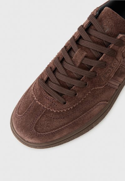Brown suede sneaker with rounded toe, textured surface, flat brown laces, and subtle branding detail near the tongue. Features a rubber sole.