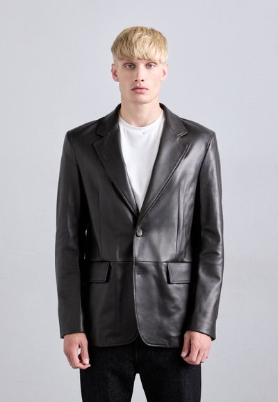 Joven con cabello rubio que lleva una chaqueta de cuero negra sobre una camiseta blanca y pantalones negros, de pie contra un fondo liso de color gris claro.