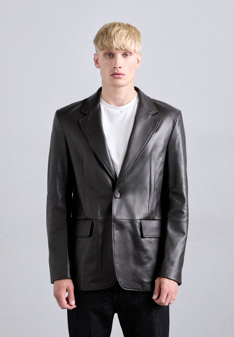 Young man with blonde hair wearing a black leather blazer over a white t-shirt and black pants, standing against a plain light gray background.