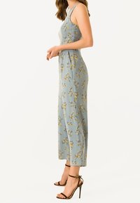 Lyseblå blomsterprintet jumpsuit med tætsiddende krop, brede ben og lommer, med gule og hvide mønstre, stylet med sorte hæle med stropper.