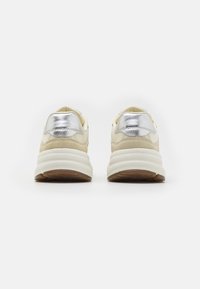 Baskets beiges avec des accents métalliques argentés et une épaisse semelle blanche, présentant une texture en daim et une surface lisse au niveau du talon.