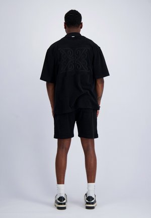Mannelijk model staat met de rug naar de camera, draagt een zwart T-shirt met korte mouwen met een subtiel rugpatroon, zwarte korte broek, witte sokken en zwart-witte sneakers.