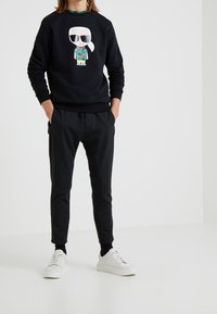 Sudadera negra con un gráfico de un personaje en una camisa de flores, combinada con pantalones tapered negros y zapatillas blancas.
