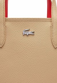 Sac à main en cuir beige avec une surface texturée, arborant un logo crocodile argenté de Lacoste et des poignées rouges.