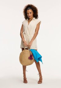 Világos bézs átkötős ruha rövid ujjakkal, deréknál húzott, texturált anyagból. Pamut szatyorral, kék törülközővel és rózsaszín bojtokkal párosítva.