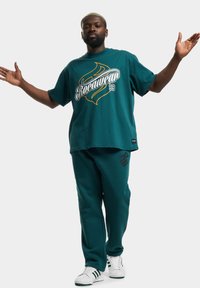 Tyrkysové oversized tričko s grafikou "Rocawear 99". Odpovídající tyrkysové tepláky s tmavým logem. Bílé sneakers s černými a zelenými akcenty.