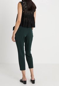 Černý krajkový top s vroubkovanými okraji v kombinaci se zelenými elegantními kalhotami. Model má na sobě černé muly na blokovém podpatku. Pohled zezadu ukazuje vrstvený design.