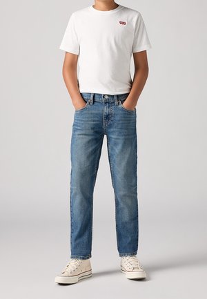 Junge Person trägt ein weißes Levi's T-Shirt, blaue Jeans und weiße Sneakers, steht mit den Händen in den Taschen vor einem einfarbigen Hintergrund.
