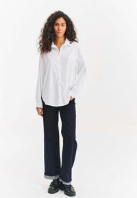 Bílá košile na knoflíky s dlouhými rukávy, zaobleným spodním lemem a přední kapsou. Kombinovaná s tmavě modrými džíny se širokými nohavicemi a černými platformovými botami.