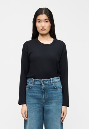 Fiatal nő hosszú, sötét hajjal, fekete hosszú ujjú pólót és magas derekú kék farmert viselve, sima világos háttér előtt áll.