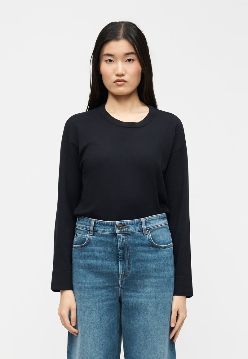 Jeune femme aux longs cheveux bruns portant un haut à manches longues noir et un jean taille haute bleu, debout devant un fond clair uni.