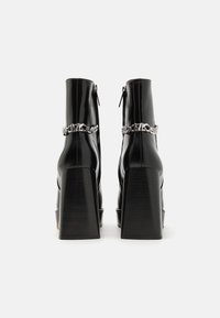 Bottines à plateforme en cuir verni noir avec un talon épais, présentant un accent en chaîne argentée autour de la cheville et une fermeture éclair au dos.