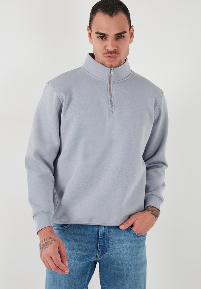 Sudadera gris claro con cuello alto y cremallera de un cuarto. Confeccionada en una tela suave, tiene mangas largas y puños de canalé, combinada con jeans azules.