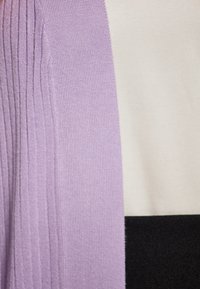 Kardigan knit dengan lengan ungu beradik, dipadukan dengan panel krim dan hitam. Detail desain tekstur lembut dan pemblokiran warna.