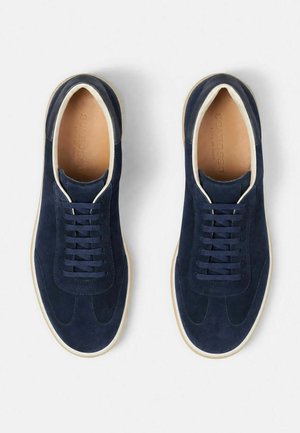 Een paar marineblauwe suède sneakers met witte biezen en veters, van bovenaf gezien op een witte achtergrond.