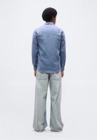 Άνδρας με αφροχτένισμα που κοιτάζει προς τα πίσω, φορώντας μπλε πουκάμισο με μακριά μανίκια και ανοιχτόχρωμα τζιν με φαρδιά μπατζάκια, σε απλό λευκό φόντο.