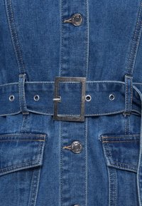 Τζιν φόρεμα σε ανοιχτό γαλάζιο χρώμα, με ζώνη που φέρει ορθογώνια ασημένια αγκράφα, κουμπιά τύπου κλιπ και τσέπες μπροστά. Η υφή είναι λεία και στιβαρή.