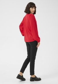 Röd långärmad blus med lös passform och samlade axlar, i kombination med svarta slim-fit byxor och svarta skor med tjocka sulor.