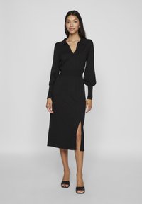 Juodas vidutinio ilgio suknelė su apykakle, ilgomis siaurėjančiomis rankovėmis, prigludusia juosmens dalimi ir šonine išpjova. Lygaus tekstūros, minimalistiško dizaino.
