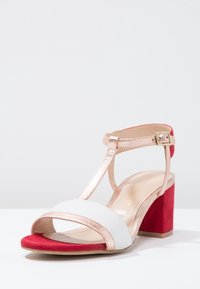 Sandale à talons hauts blanche et rouge avec un talon carré, des brides en métal rose doré et une texture douce en suède sur le talon et la semelle.