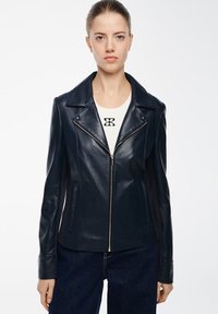 Giacca da biker in pelle blu navy con zip frontale, ampie alette e cuciture laterali. Indossata sopra una maglietta bianca con un logo nero, abbinata a jeans scuri.