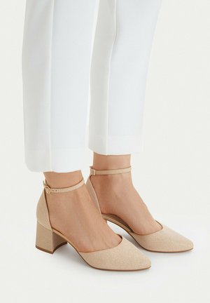 Zapatos beige con brillo, tacón de bloque y correa en el tobillo, usados con pantalones blancos cortos sobre un fondo claro.