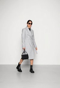 Ljusgrå knälång kappa med skärp i midjan, sjalkrage och långa ärmar, matchad med svarta ankelboots och en liten handväska.