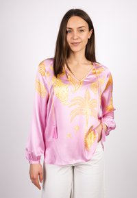 Rosa Satinbluse mit goldfarbenen Mustern, darunter Tiere und Palmen. Verfügt über einen V-Ausschnitt und Quasten am Ausschnitt sowie an den Ärmeln.