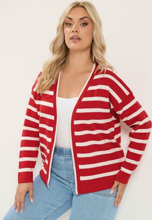 Vrouw draagt een rood-wit horizontaal gestreepte cardigan, wit topje en lichtblauwe spijkerbroek, staand met één hand op haar heup.
