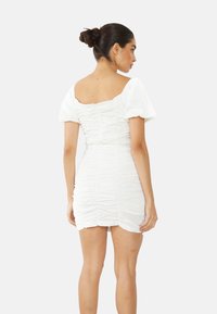 Mujer con cabello oscuro recogido en un moño, vestida con un vestido corto y fruncido de color blanco con mangas abullonadas, de pie y de espaldas sobre un fondo liso.
