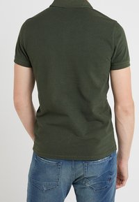 Homme portant un polo à manches courtes vert foncé et un jean bleu, vu de dos sur un fond clair et uni.