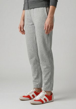 Osoba v šedých teplácích a červenobílých teniskách stojí před jednobarevným světlým pozadím.