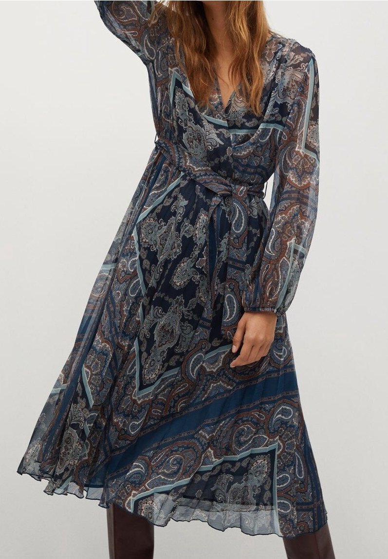 Femme portant une robe portefeuille à manches longues bleu marine à motif cachemire, ceinture nouée à la taille, jupe fluide et bottes marron montant au genou.