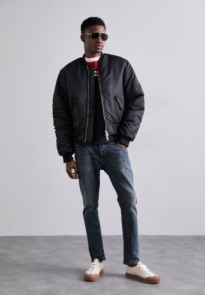 Giacca bomber nera, maglione grigio con dettagli rossi e bianchi, jeans blu, sneakers chiare con suole in gomma e occhiali da sole. Posizione in piedi.