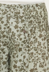 Slap-fit bukser i lysegrønt stof med brune blomstertryk. Udstyret med rynkede detaljer langs taljen og en lille dekorativ charme.