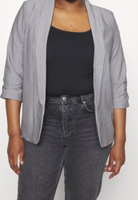 Grauer Blazer mit weicher Textur und offenem Design, kombiniert mit einem schwarzen Oberteil und dunklen Jeans, mit hochgerollten Ärmeln und subtiler Schnittführung.