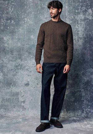 Pull en tricot marron avec un motif texturé, associé à un jean foncé et des chaussures marron. L'arrière-plan est d'un gris atténué.