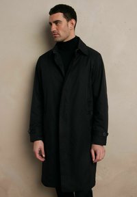 Czarny płaszcz trench z zapięciem na guziki przy kołnierzu i mankietach, charakteryzujący się eleganckim fasonem. Materiał ma gładkie wykończenie i sięga do połowy uda.