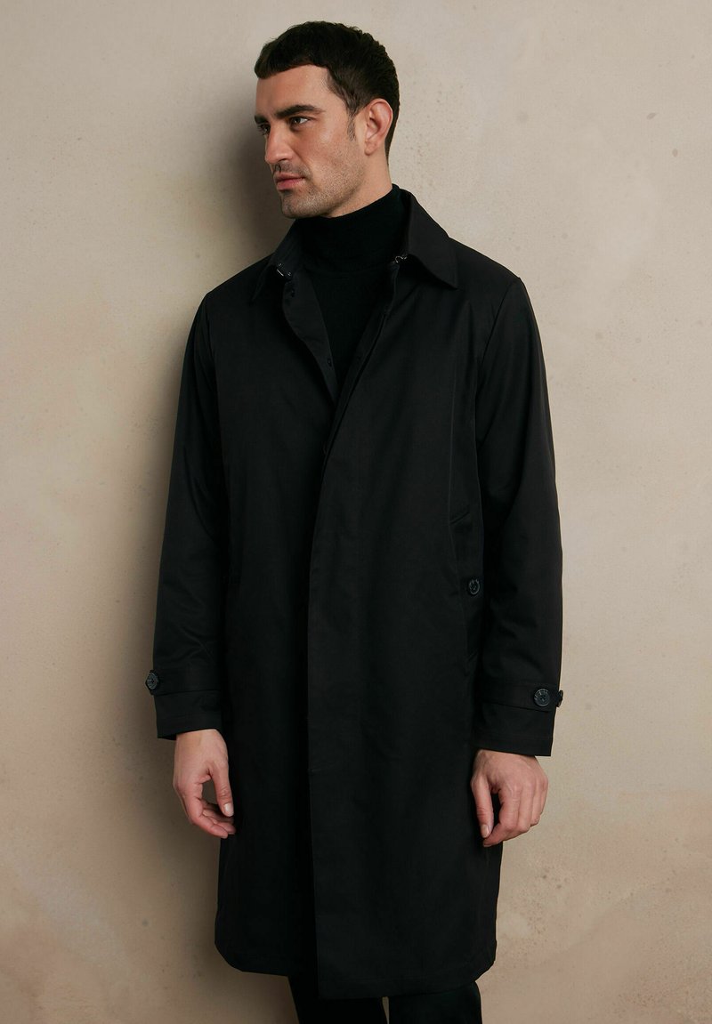 Czarny płaszcz trench z zapięciem na guziki przy kołnierzu i mankietach, charakteryzujący się eleganckim fasonem. Materiał ma gładkie wykończenie i sięga do połowy uda.