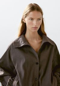 Camicia marrone con bottoni e ampio colletto, realizzata in tessuto testurizzato. Presenta bottoni frontali e maniche leggermente oversize.