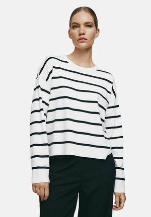 Giovane donna con i capelli legati indietro, che indossa un maglione bianco a maniche lunghe con strisce orizzontali nere e pantaloni neri, in piedi contro uno sfondo bianco.