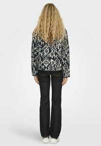 Giacca a maglia con motivo in navarre e crema con un design geometrico, abbinata a jeans scuri a zampa d'elefante e sneakers bianche. Vista dalla parte posteriore.
