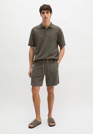Junger Mann steht und trägt ein strukturiertes olivgrünes Poloshirt und passende Shorts mit Kordelzug sowie graue Slip-On-Sandalen, neutraler Hintergrund.